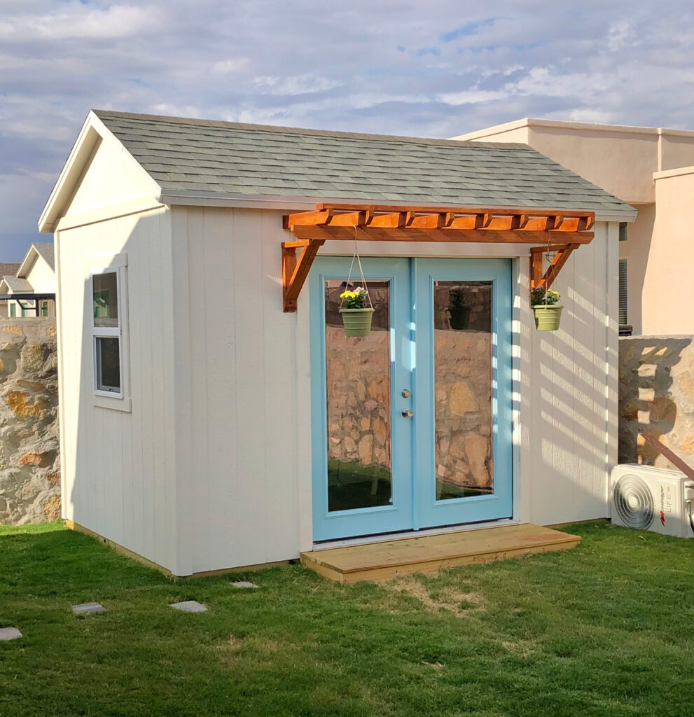 A white shed with blue doors and flower pots hanging from the front in an El Paso backyard.